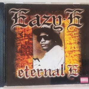 Op de afbeelding: Een compact disc hoesje met het album Eternal E van Eazy-E. De hoes toont een portret van Eazy-E met een zonnebril en een pet, gecentreerd tegen een vurige oranje en zwarte achtergrond met de tekst Eazy-E en eternal E.