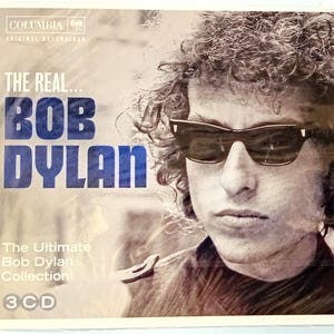 May include: A 3-CD set of Bob Dylan music. The cover features a black and white photo of Bob Dylan wearing sunglasses. The text on the cover reads "The Real... Bob Dylan" and "The Ultimate Bob Dylan Collection".