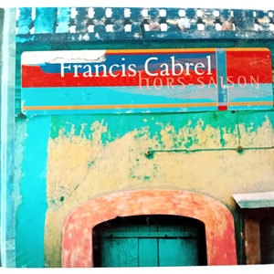 Puede incluir: Un edificio turquesa y amarillo con una etiqueta roja y amarilla que dice "Francis Cabrel Hors Saison".