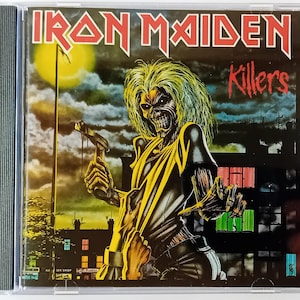 May include: A CD case with a black background and a yellow and black image of a man with a skull face holding a hammer. The text "Iron Maiden" and "Killers" is printed on the case.
