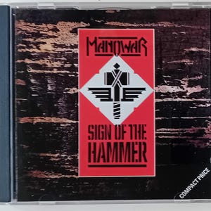 May include: A red CD case with a black and white image of a hammer and the text "Manowar Sign of the Hammer" on the front. The case also has the text "Compact Price" in the bottom right corner.