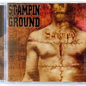 Op de afbeelding: Een CD-hoes met een grunge-achtige afbeelding van een persoon met gestikte huid. De tekst "Stampin' Ground" staat in zwarte letters op een rode achtergrond. Het woord "Sacred" staat in rood op de borst van de persoon.