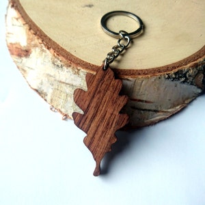 Könnte beinhalten: Ein Schlüsselanhänger aus Holz in Form eines Eichenblattes mit einem silbernen Schlüsselring. Das Blatt ist dunkelbraun und hat eine natürliche Holzmaserung.