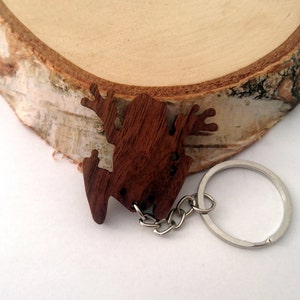 Peut inclure: Un porte-clés en forme de grenouille en bois avec un anneau et une chaîne argentés. La grenouille est brune et présente un design détaillé.
