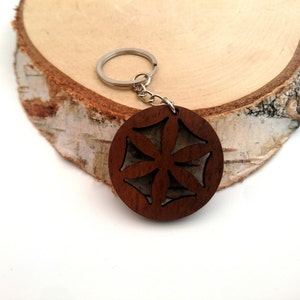 Puede incluir: Un llavero redondo de madera con un diseño floral recortado. El llavero es marrón y está unido a un anillo de metal plateado. El fondo es una rodaja de madera de color claro.