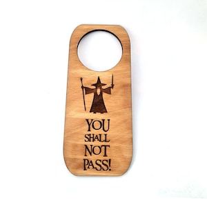 Può includere: Appendiporta in legno con una silhouette di Gandalf de Il Signore degli Anelli. Il testo "YOU SHALL NOT PASS!" è inciso sull'appendiporta.