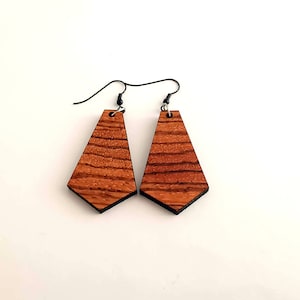 Può includere: Un paio di orecchini in legno con forma geometrica. Gli orecchini sono in legno marrone scuro con un motivo a venature naturali. Gli orecchini hanno un gancio argentato.