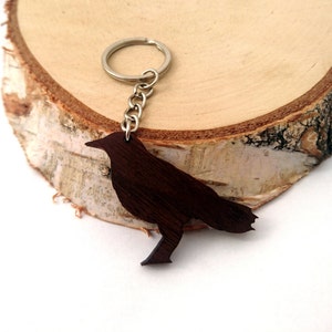 May include: A dark brown wooden crow keyring with a silver chain and ring. The keyring is sitting on a piece of wood.