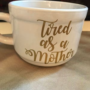 May include: White ceramic mug with a gold inscription that reads "Tired as a Mother".