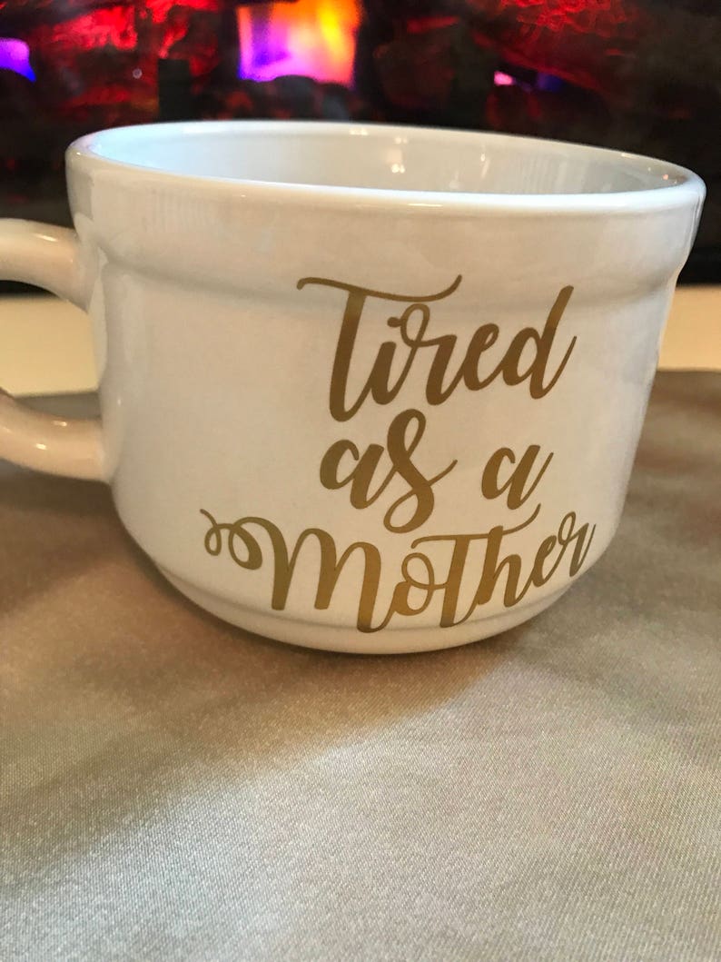 May include: White ceramic mug with a gold inscription that reads "Tired as a Mother".