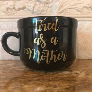 May include: Black ceramic mug with a gold inscription that reads "Tired as a Mother".