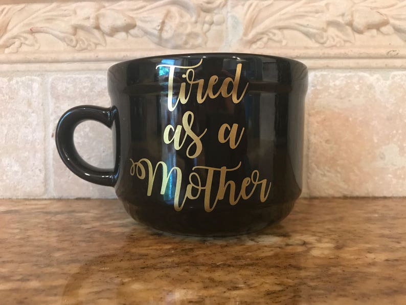 May include: Black ceramic mug with a gold inscription that reads "Tired as a Mother".