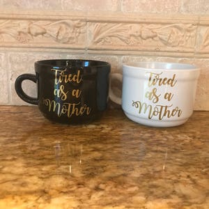 May include: Two ceramic mugs with the text "Tired as a Mother" in gold lettering. One mug is black and the other is white.