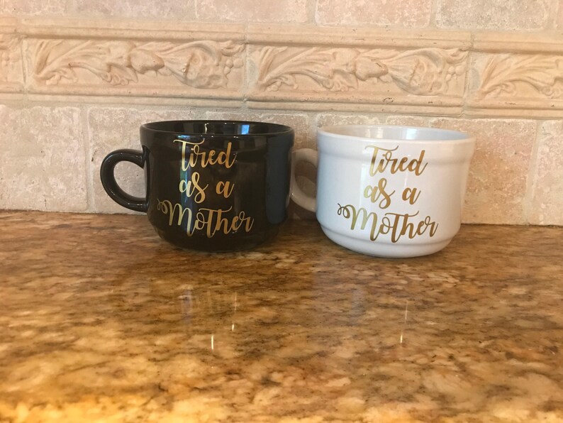 May include: Two ceramic mugs with the text "Tired as a Mother" in gold lettering. One mug is black and the other is white.