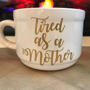 May include: White ceramic mug with a gold inscription that reads "Tired as a Mother".