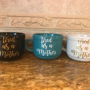 May include: Three ceramic mugs with the text "Tired as a Mother" in gold lettering. The mugs are black, teal, and white.