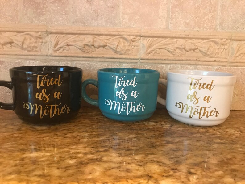 May include: Three ceramic mugs with the text "Tired as a Mother" in gold lettering. The mugs are black, teal, and white.