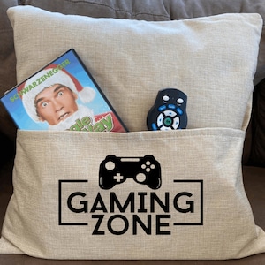 May include: A beige pillow with a pocket on the front. The pocket contains a DVD and a remote control. The pillow has black text that says "GAMING ZONE" and a black video game controller graphic.