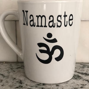 May include: White ceramic mug with a black inscription that reads "Namaste" and a black Om symbol.