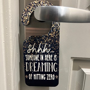 May include: A black door hanger with gold glitter accents. The hanger has a white star on each side of the text, "Shhh... Someone in here is dreaming of hitting zero."