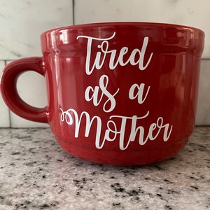 May include: Red ceramic coffee mug with a white inscription that reads "Tired as a Mother".