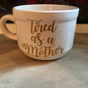 May include: White ceramic mug with a gold inscription that reads "Tired as a Mother".