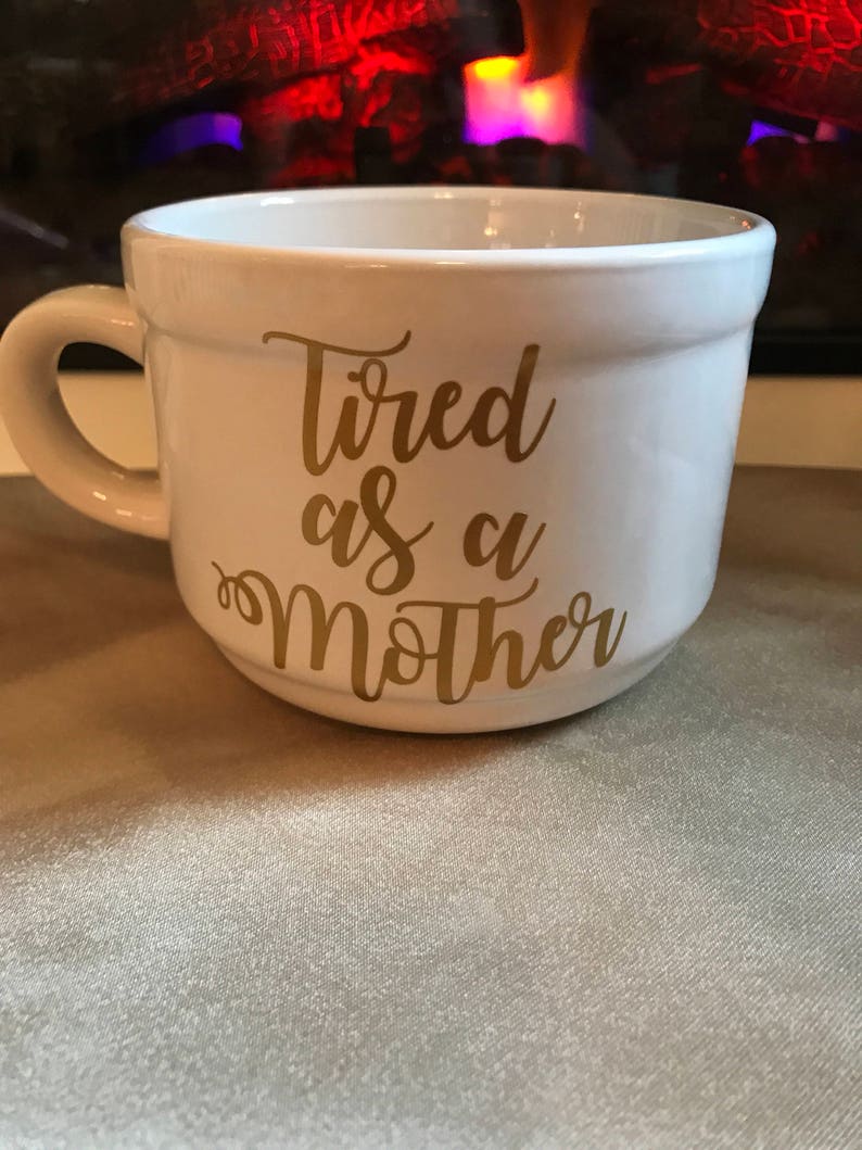 May include: White ceramic mug with a gold inscription that reads "Tired as a Mother".
