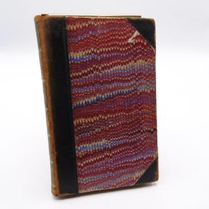 Könnte beinhalten: Ein Vintage-Buch mit einem rot-weiß-blau marmorierten Einband. Das Buch hat einen schwarzen Rücken und ist in Leder gebunden.