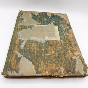 Puede incluir: Un libro antiguo de tapa dura, verde y marrón, con una cubierta desgastada y un trozo de papel pegado en la parte delantera.