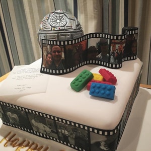 K&ouml;nnte beinhalten: Ein wei&szlig;er Kuchen, der mit einem Filmstreifenrand und der Aufschrift "Happy Birthday" verziert ist. Der Kuchen ist mit einem silbernen Todesstern, Lego-Steinen und einer handgeschriebenen Notiz verziert.