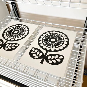 May include: Two black and white prints of stylized flowers with a circular design. The prints are on white paper and are drying on a white metal rack.