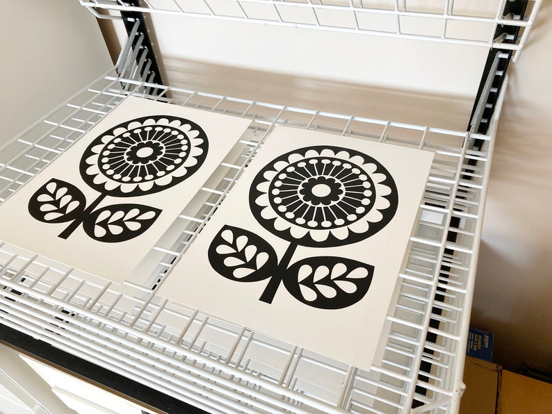 May include: Two black and white prints of stylized flowers with a circular design. The prints are on white paper and are drying on a white metal rack.