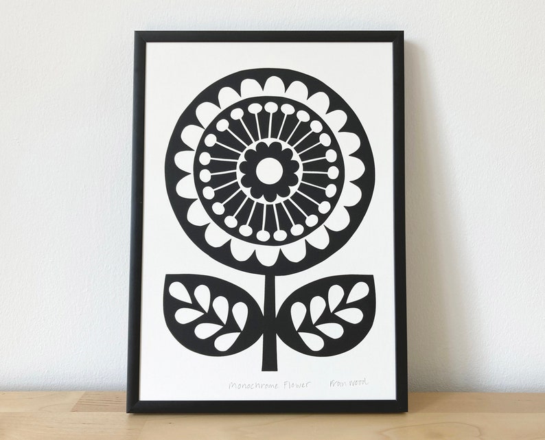 May include: Black and white print of a stylised flower with a large circular centre and two leaves. The print is framed in a black frame and has the text "Monochrome Flower" and "from paper" written below the image.