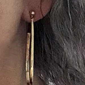 May include: A pair of gold hoop earrings with a slightly hammered texture. The earrings are worn in a person's ear.