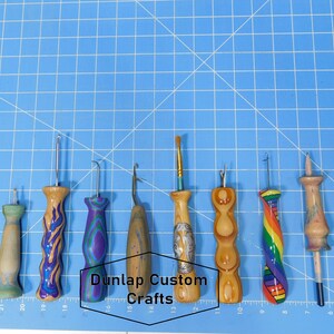 Puede incluir: Un conjunto de 10 ganchillos de madera con mangos de diferentes colores y tama&ntilde;os de gancho. Los ganchillos est&aacute;n dispuestos en una fila sobre una superficie azul cuadriculada. El texto "Dunlap Custom Crafts" es visible en el centro de la imagen.