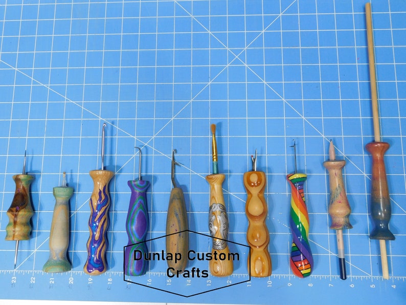 Puede incluir: Un conjunto de 10 ganchillos de madera con mangos de diferentes colores y tama&ntilde;os de gancho. Los ganchillos est&aacute;n dispuestos en una fila sobre una superficie azul cuadriculada. El texto "Dunlap Custom Crafts" es visible en el centro de la imagen.