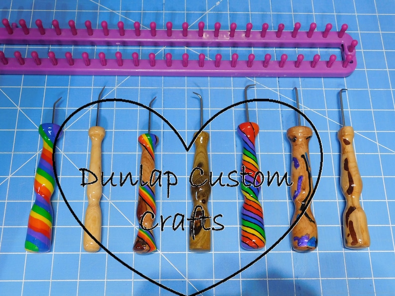 Peut inclure: Un m&eacute;tier &agrave; tricoter en plastique violet et sept crochets en m&eacute;tal avec des manches d&eacute;coratifs en bois et aux motifs arc-en-ciel, avec le texte Dunlap Custom Crafts dans un c&oelig;ur noir sur un tapis de d&eacute;coupe quadrill&eacute; bleu.