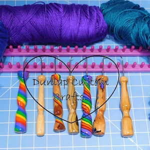 May include: Six loom knitting hooks with decorative handles featuring rainbow patterns and natural wood grains, arranged in front of a purple plastic knitting loom and three skeins of blue, purple, and teal yarn on a blue cutting mat.