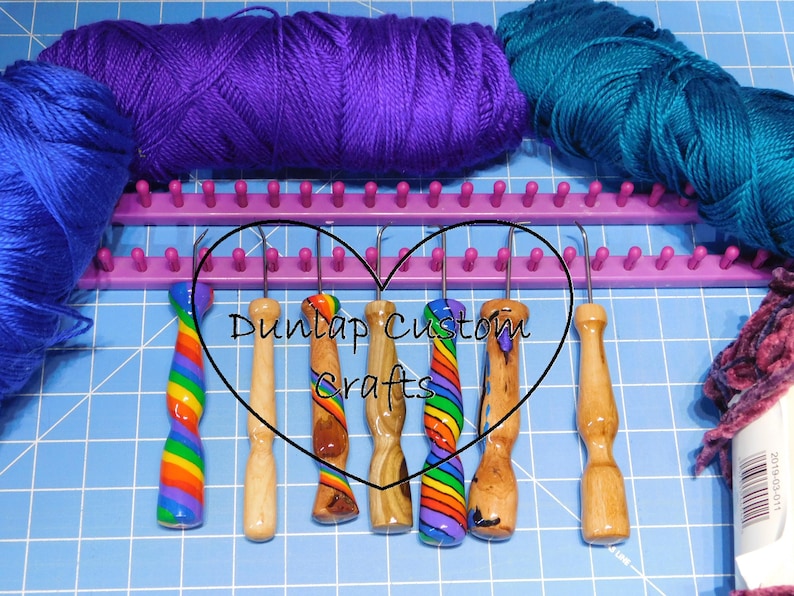 Peut inclure: Six crochets &agrave; tricoter avec des manches d&eacute;coratifs aux motifs arc-en-ciel et aux grains de bois naturels, dispos&eacute;s devant un m&eacute;tier &agrave; tricoter en plastique violet et trois &eacute;cheveaux de laine bleue, violette et sarcelle sur un tapis de coupe bleu.