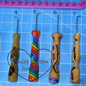 Peut inclure: Cinq crochets de m&eacute;tier &agrave; tisser en m&eacute;tal avec des manches en bois tourn&eacute;, num&eacute;rot&eacute;s de 3 &agrave; 7, pr&eacute;sentant divers grains de bois et motifs arc-en-ciel. Un cadre de m&eacute;tier &agrave; tisser violet est visible en haut. Le texte Dunlap Custom Crafts est superpos&eacute;.