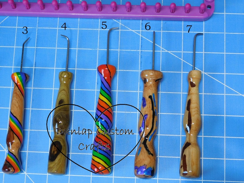 Peut inclure: Cinq crochets de m&eacute;tier &agrave; tisser en m&eacute;tal avec des manches en bois tourn&eacute;, num&eacute;rot&eacute;s de 3 &agrave; 7, pr&eacute;sentant divers grains de bois et motifs arc-en-ciel. Un cadre de m&eacute;tier &agrave; tisser violet est visible en haut. Le texte Dunlap Custom Crafts est superpos&eacute;.