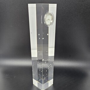 May include: A clear acrylic clock with a white face and black hands. The clock is encased in a tall, rectangular block of acrylic with small, clear bubbles throughout.