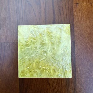 Puede incluir: Un azulejo cuadrado de mármol amarillo y blanco con un patrón de remolino. El azulejo está hecho de un material translúcido y tiene un acabado brillante.