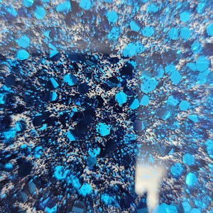 May include: Abstract close-up of a surface with a textured, speckled appearance. The surface is predominantly blue, with varying shades of turquoise and navy. Irregular, geometric shapes and small particles create a dynamic, visually engaging pattern.