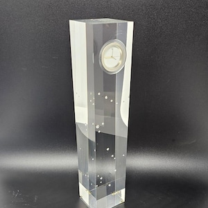 Może przedstawiać: Przezroczysty zegar akrylowy z białą tarczą i czarnymi wskazówkami. Zegar jest osadzony w wysokim, prostokątnym bloku akrylowym z małymi, przezroczystymi bąbelkami.