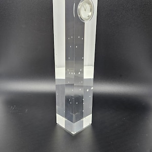 May include: A clear, rectangular clock with a silver face and hands. The clock is encased in a clear, geometric block with white vertical stripes. The block has a faceted base and is set against a dark background.