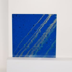 Puede incluir: Una pieza de arte abstracto cuadrada azul y blanca con una textura brillante. El fondo azul tiene rayas blancas de pintura y purpurina iridiscente.