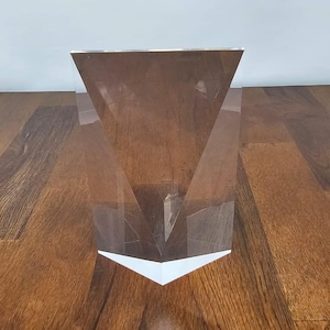 Könnte beinhalten: Eine klare, geometrische Acrylskulptur mit dreieckiger Oberseite und V-förmiger Basis. Die Skulptur ist transparent, lässt Licht durch und wird auf einer Holzoberfläche präsentiert.