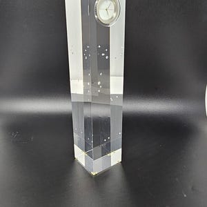 May include: A clear acrylic clock with a white face and black hands. The clock is encased in a rectangular block of clear acrylic with small, clear, round accents throughout.