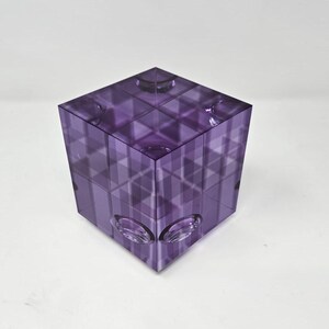 Peut inclure: Un cube en verre violet avec un motif géométrique. Le cube est en verre transparent avec une teinte violette. Le cube a un motif de grille sur chaque face.
